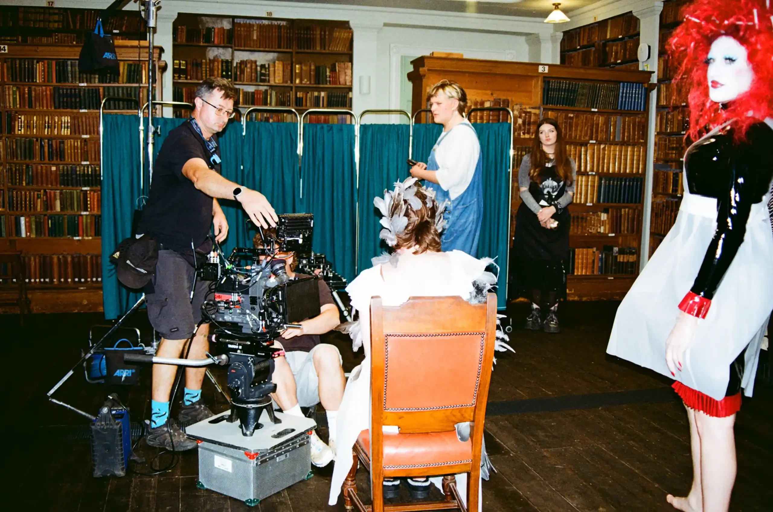 Behind the scenes of Featherless, photographed on film. A camera crew surrounds a seated performer with feathers in their hair. Teal medical screens stand between bookshelves, while another performer waits nearby in a vivid red wig, shiny black outfit and white apron.
