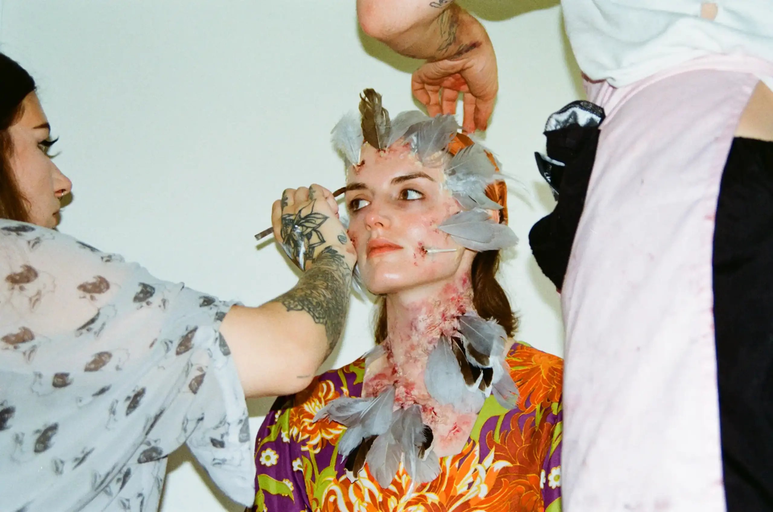 Behind the scenes of Featherless, photographed on film. Makeup artists work on a seated performer, applying feathers and reddened prosthetic skin around their face and neck. The performer looks upwards, wearing a bright orange and purple floral robe.