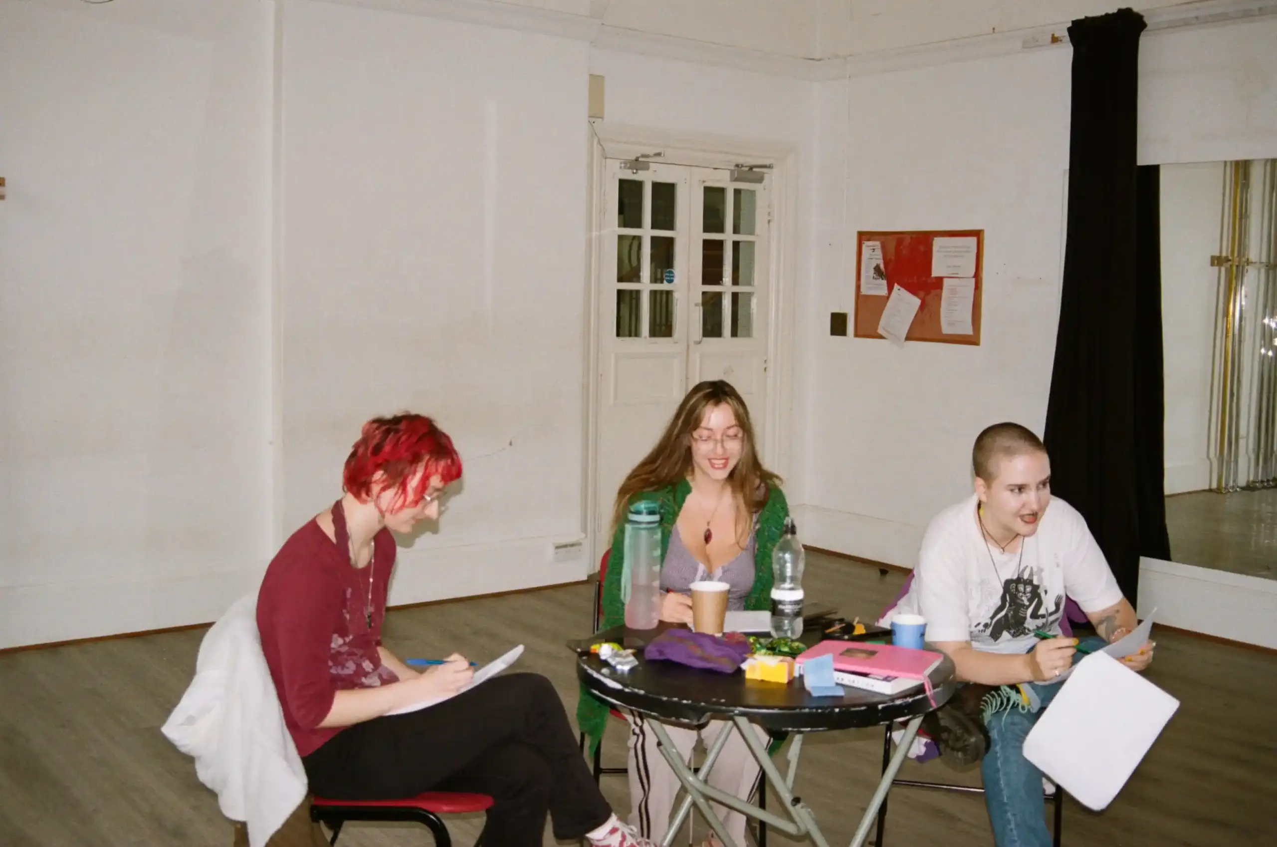 Behind the scenes of Teddy Midnight, photographed on film. Three people in their 20s sit around a small table covered with notebooks, script pages and drinks in a bright rehearsal room. One with vivid pink hair writes notes, another with long blonde hair smiles while looking down, and a third with a shaved head holds loose script pages and is in the middle of explaining something.