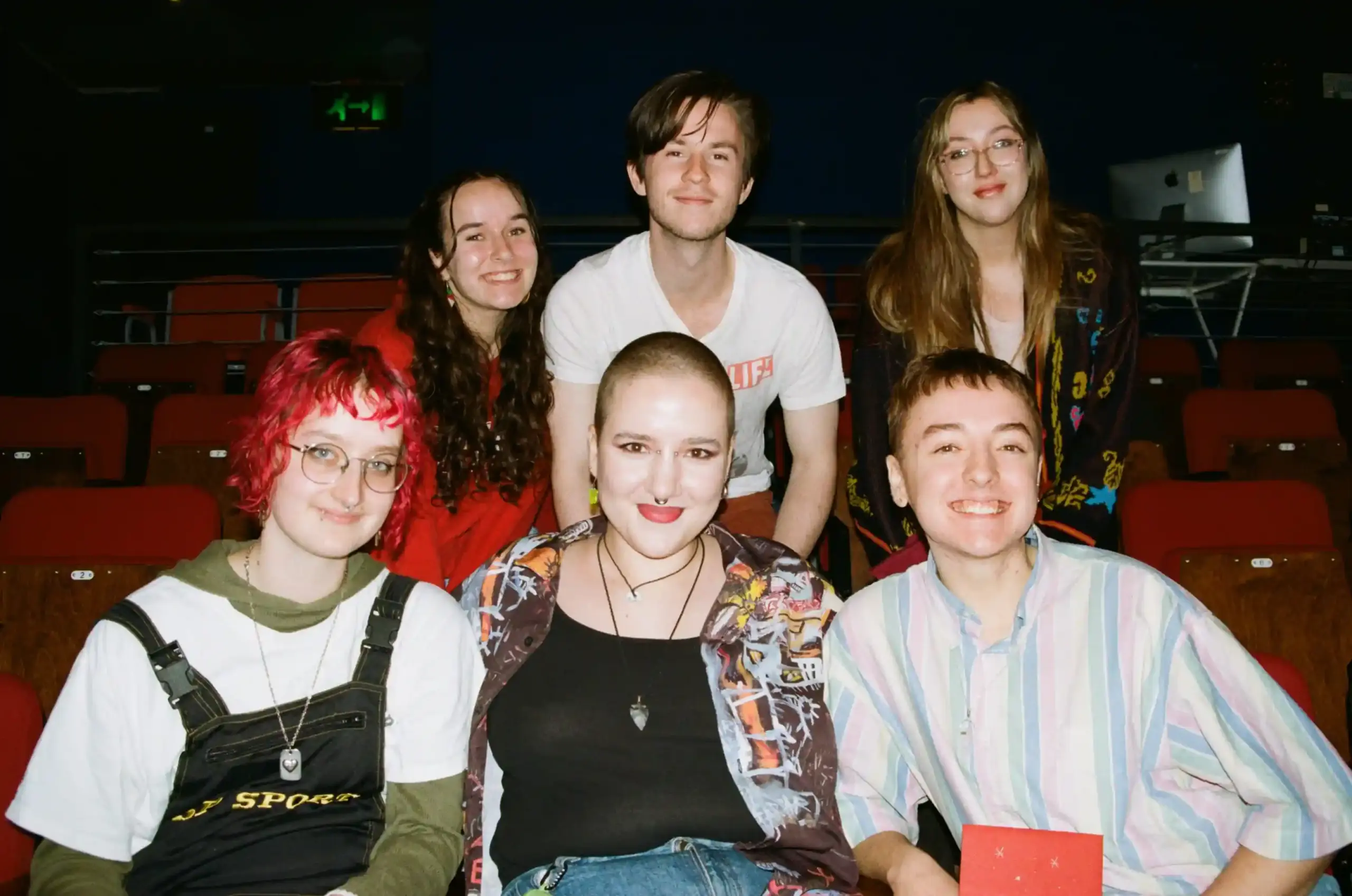 The Teddy Midnight team, people in their early 20s, photographed on film. Six people smile at the camera among rows of red theatre seats, three seated at the front and three leaning in behind them. They wear colourful casual clothes, including dungarees, patterned shirts and a red hoodie.
