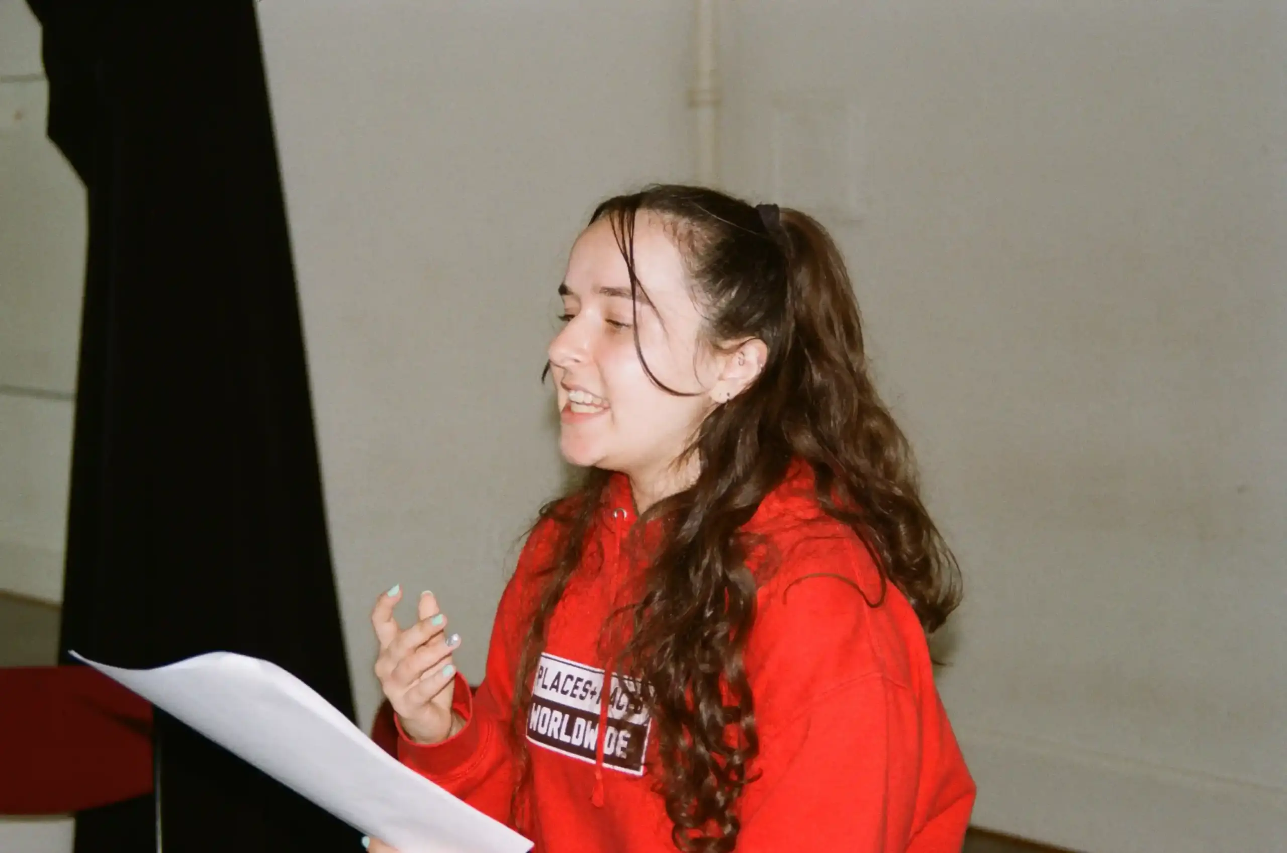 A Teddy Midnight rehearsal, photographed on film. A performer with long brown hair tied in a ponytail wears a bright red hoodie, holding script pages and gesturing with their free hand as they speak.