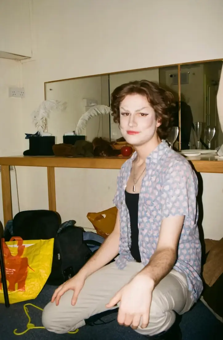 Behind the scenes of Moles, photographed on film. A person with short brown hair and dramatic eye makeup crouches in a dressing room, wearing an open blue patterned shirt, black top and beige trousers. A mirror and a counter with costume accessories are behind them.