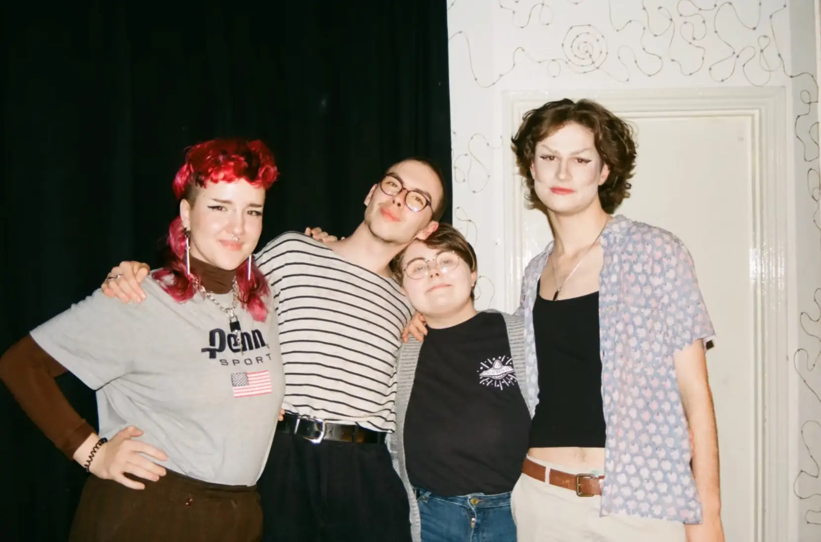 Behind the scenes of Moles, photographed on film. Four students in their early 20s pose close together with their arms around one another, against a black curtain and a white wall. One has bright red hair; another wears dramatic eye makeup and an open blue patterned shirt. Another wears a striped shirt and glasses; the fourth, a black tee and grey cardigan. 