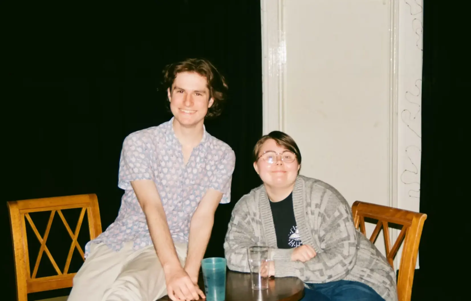 Behind the scenes of Moles, photographed on film. Two people smile at the camera beside a small round table, one wearing a blue patterned shirt and the other glasses and a grey cardigan. A black curtain hangs behind them.