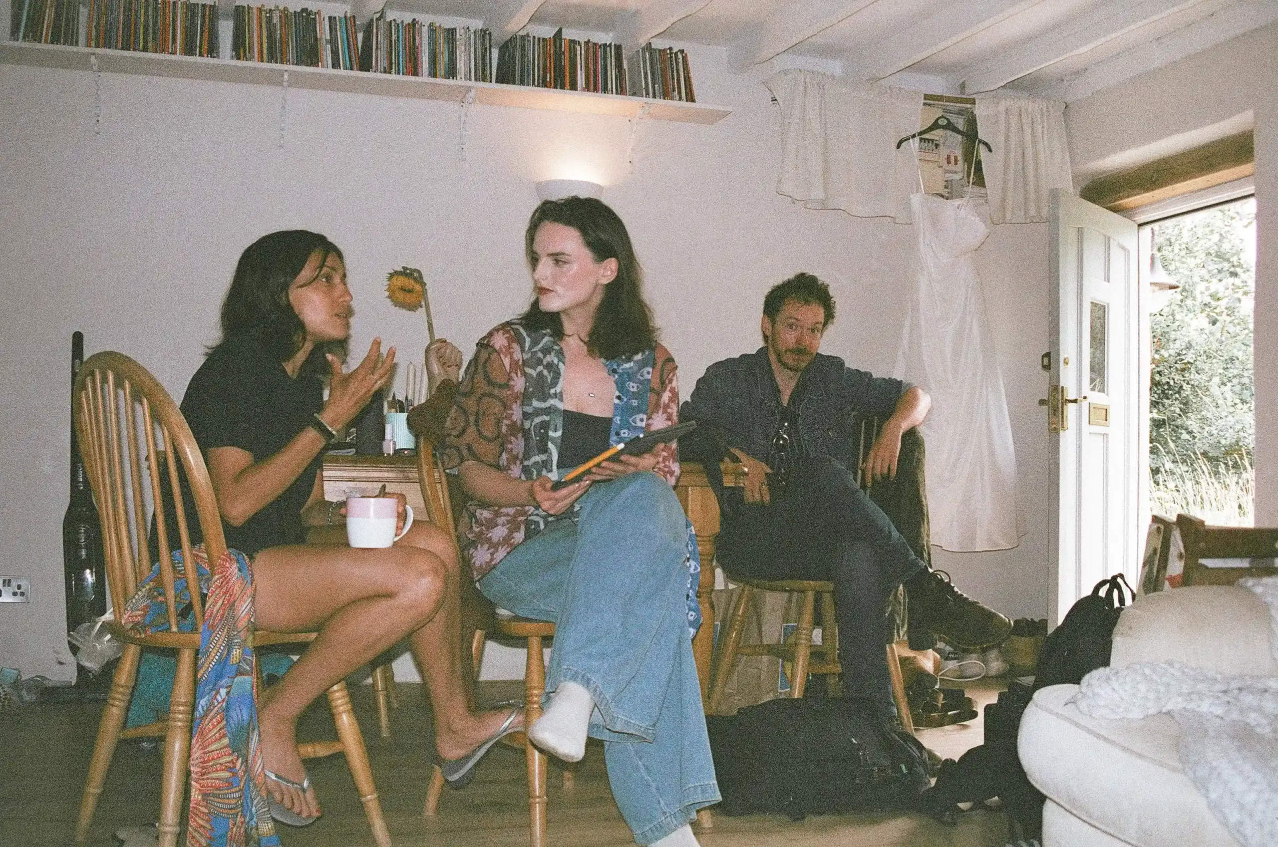 Behind the scenes of Swan, photographed on film. Three people sit in a small, bright room beneath a shelf of books. One woman in her early 30s gestures while holding a mug, another in her 20s listens in a colourful patterned shirt and jeans. A man sits beside the open doorway, looking into the camera with an expression of mock surprise, bags by his feet.