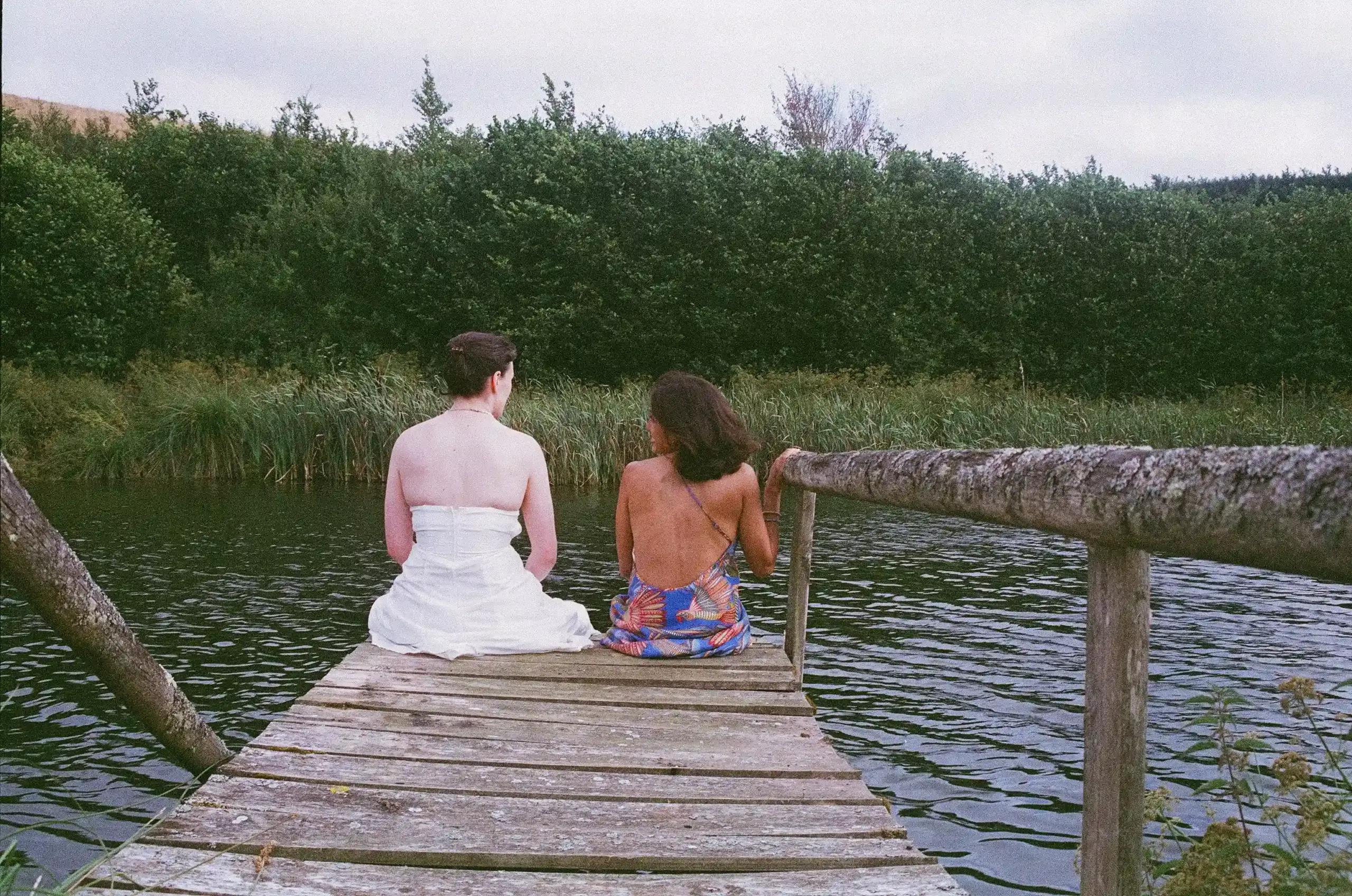 Behind the scenes of Swan, photographed on film. Two people sit beside each other at the end of a weathered wooden jetty, their backs to the camera. One wears a  strapless wedding dress and the other a colourful backless dress. Reeds and dense greenery surround the water.