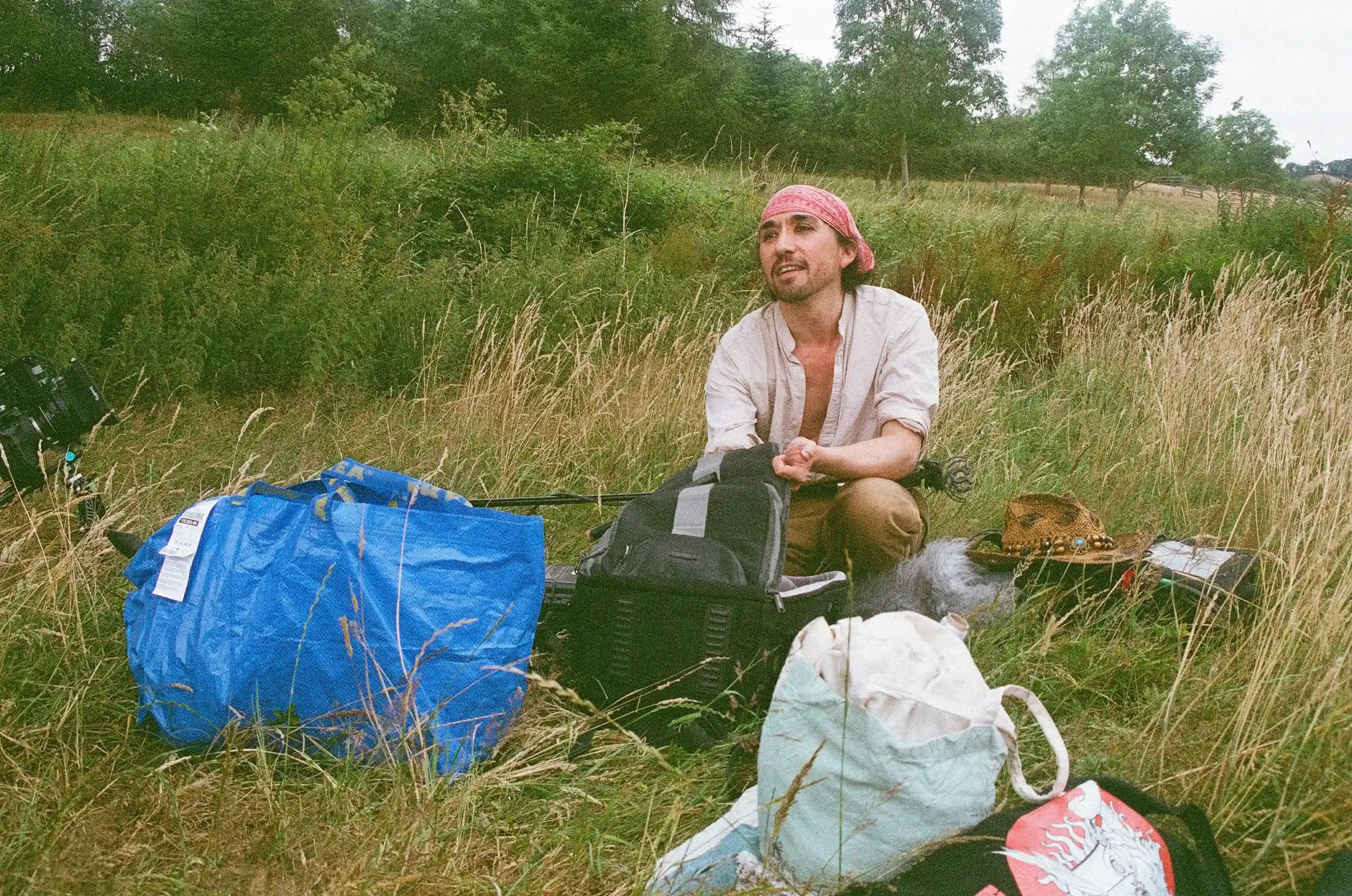 Behind the scenes of Swan, photographed on film. A person in his early 40s wearing a pink bandana and an open pale shirt sits among equipment bags in tall grass. A microphone with a furry windshield rests beside them, with a green hillside and trees beyond.